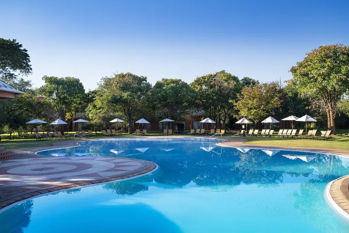The pool with sun umbrellas under shady trees at Avani Victoria Falls Resort