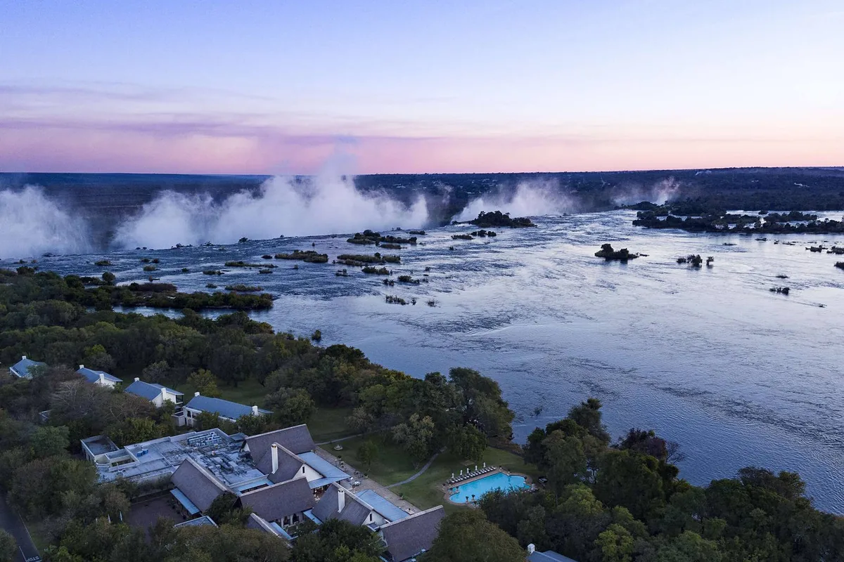 The Zambian bank of the Zambezi at dusk, with the spray of the falls rising behind the riverside lodges