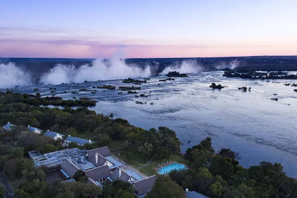 The Zambian bank of the Zambezi at dusk, with the spray of the falls rising behind the Anantara Royal Livingstone