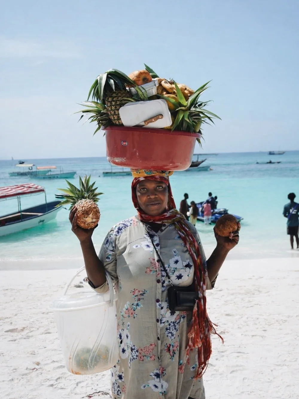 A woman carrying a basket of fruit on her head along a white-sand beach on Zanzibar